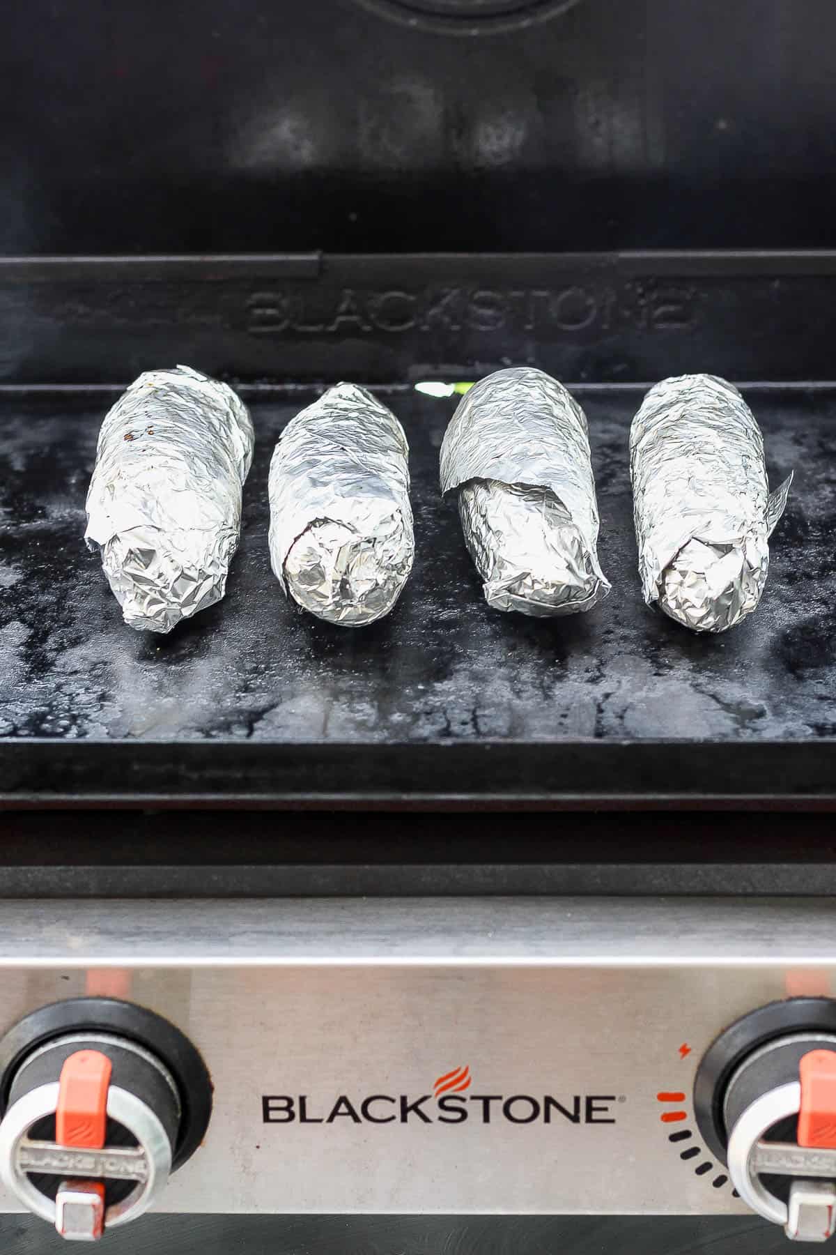Four ears of Blackstone corn on the cob, wrapped in foil, cooking on the griddle.