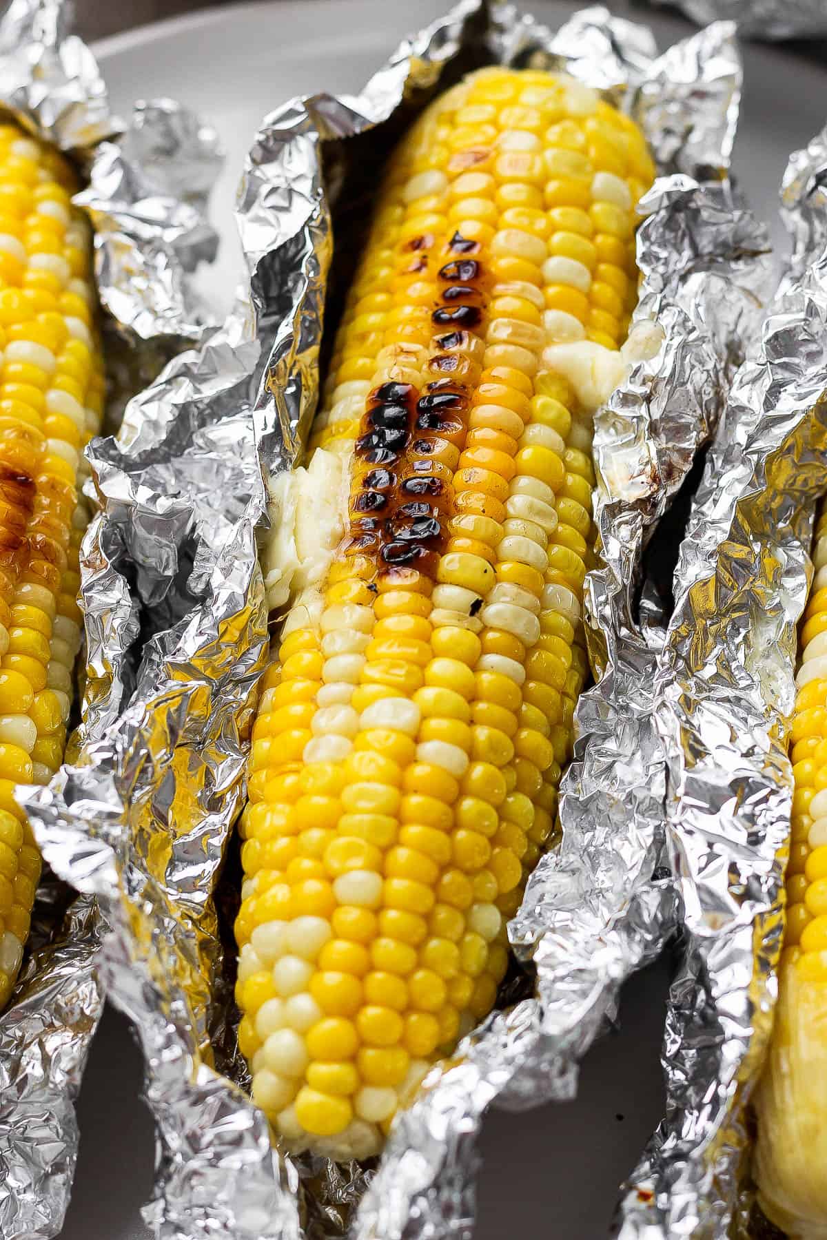 A Blackstone corn on the cob with the foil unwrapped between two other ears of corn on a plate.