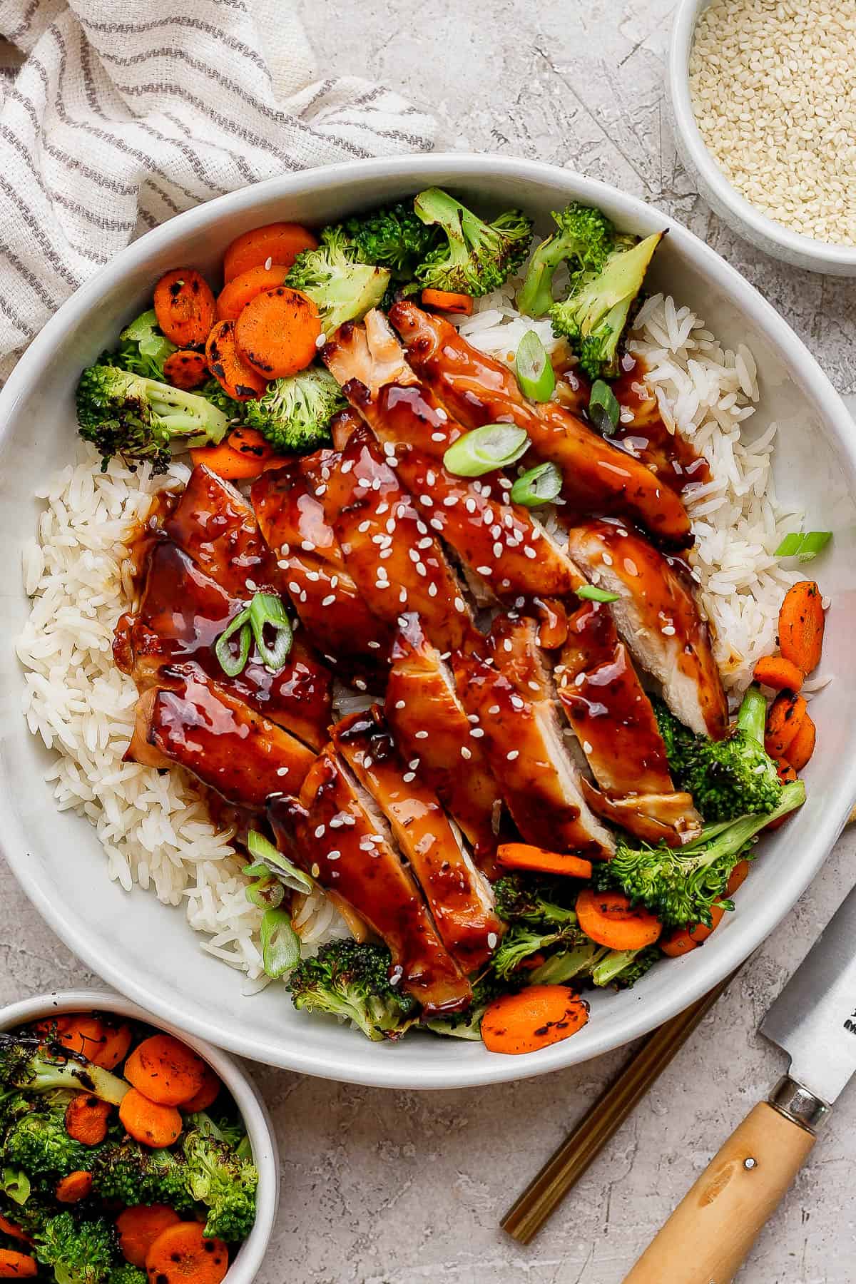 Blackstone teriyaki chicken over rice in a white bowl next to small bowls of cooked broccoli and carrots and sesame seeds.