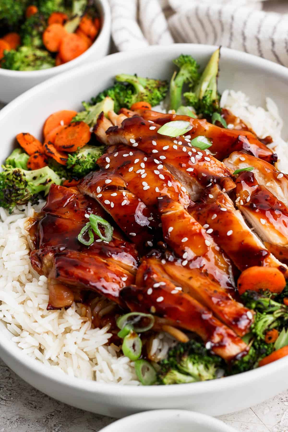 Blackstone teriyaki chicken over rice in a white bowl next to a small bowl of cooked broccoli and carrots.