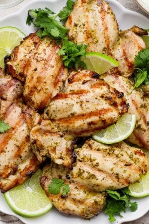 Several cilantro lime chicken thighs on a platter with lime wedges and fresh cilantro.