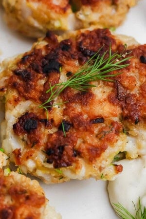 A cooked cod cake a plate with fresh dill on top and tartar sauce next to it.