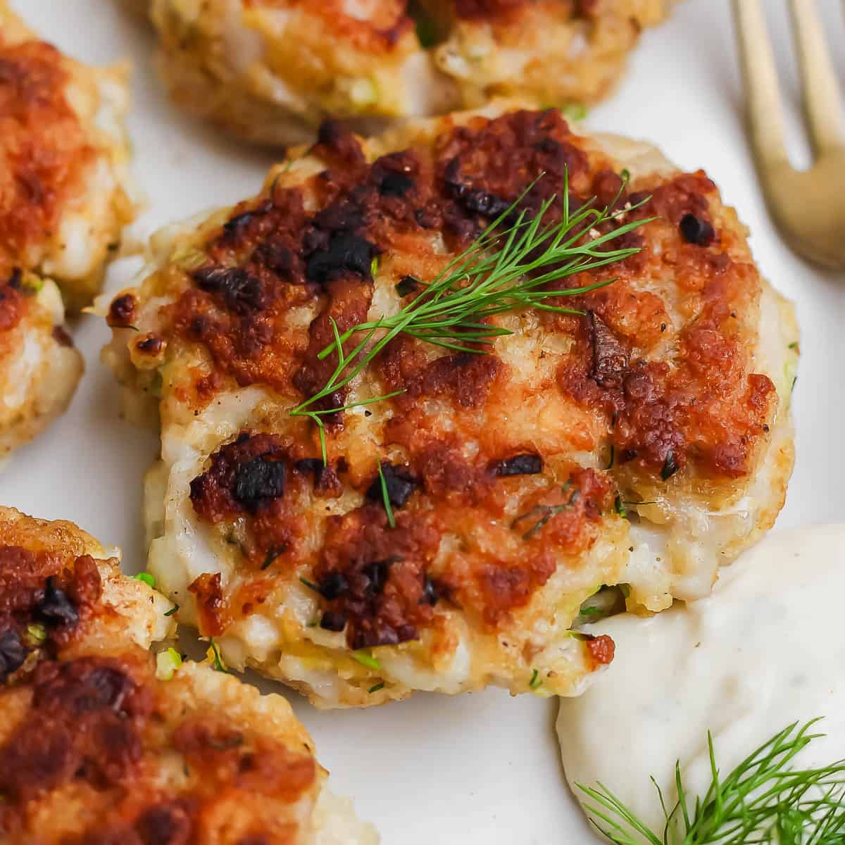A cooked cod cake a plate with fresh dill on top and tartar sauce next to it.
