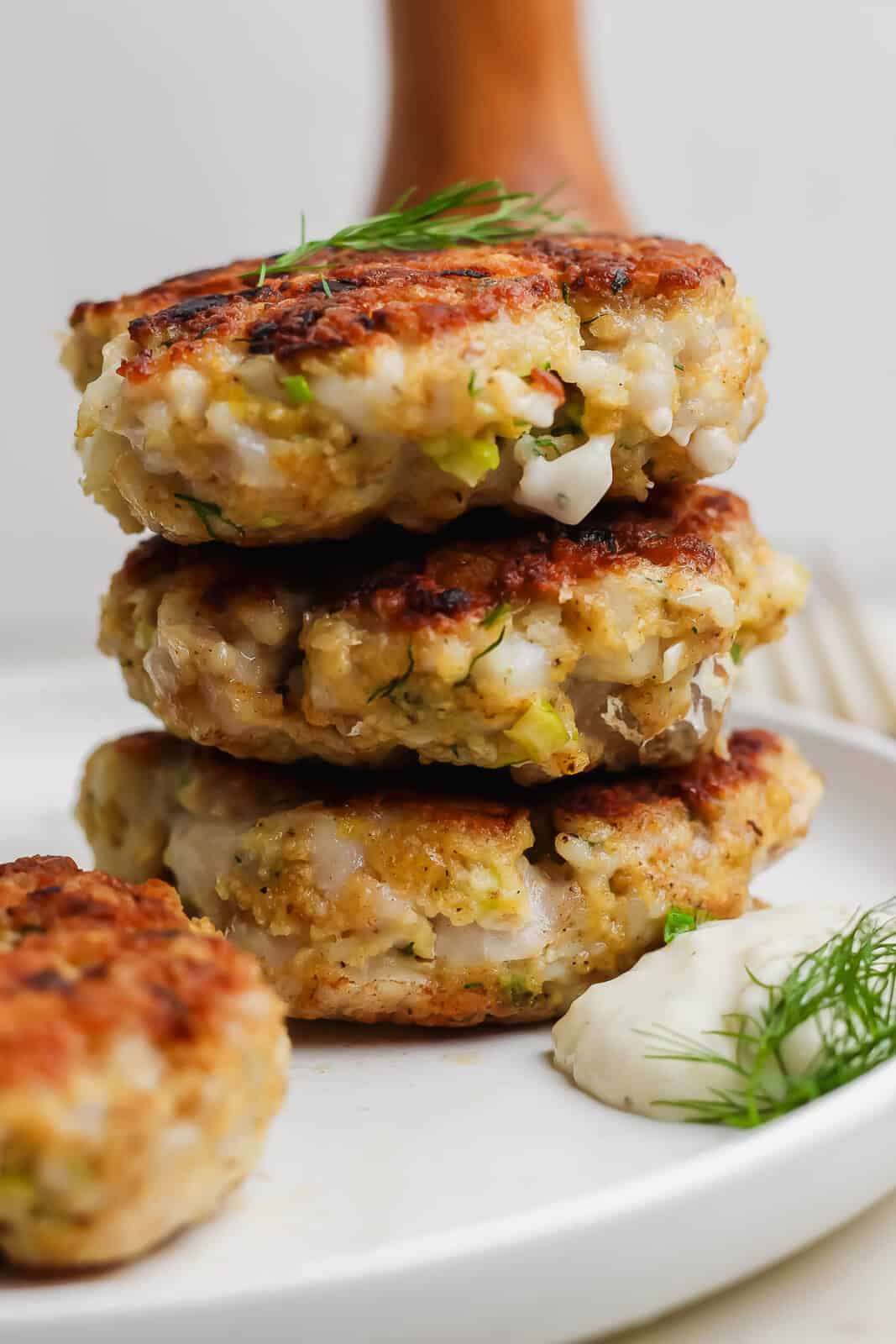 A stack of three cooked cod fish cakes on a plate next to some fresh dill and tartar sauce.