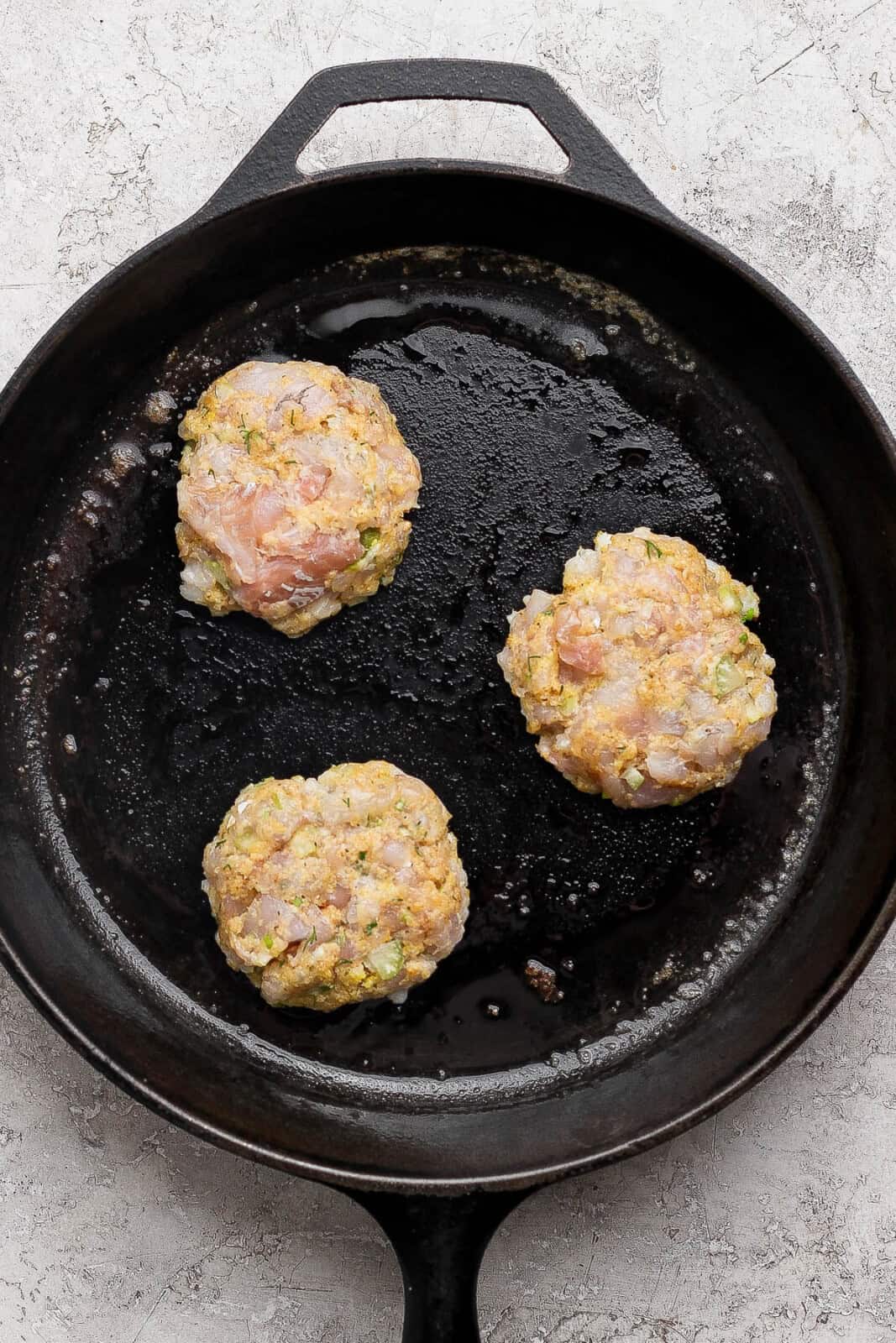Cod cakes being cooked in a cast iron skillet, but still raw on top. 