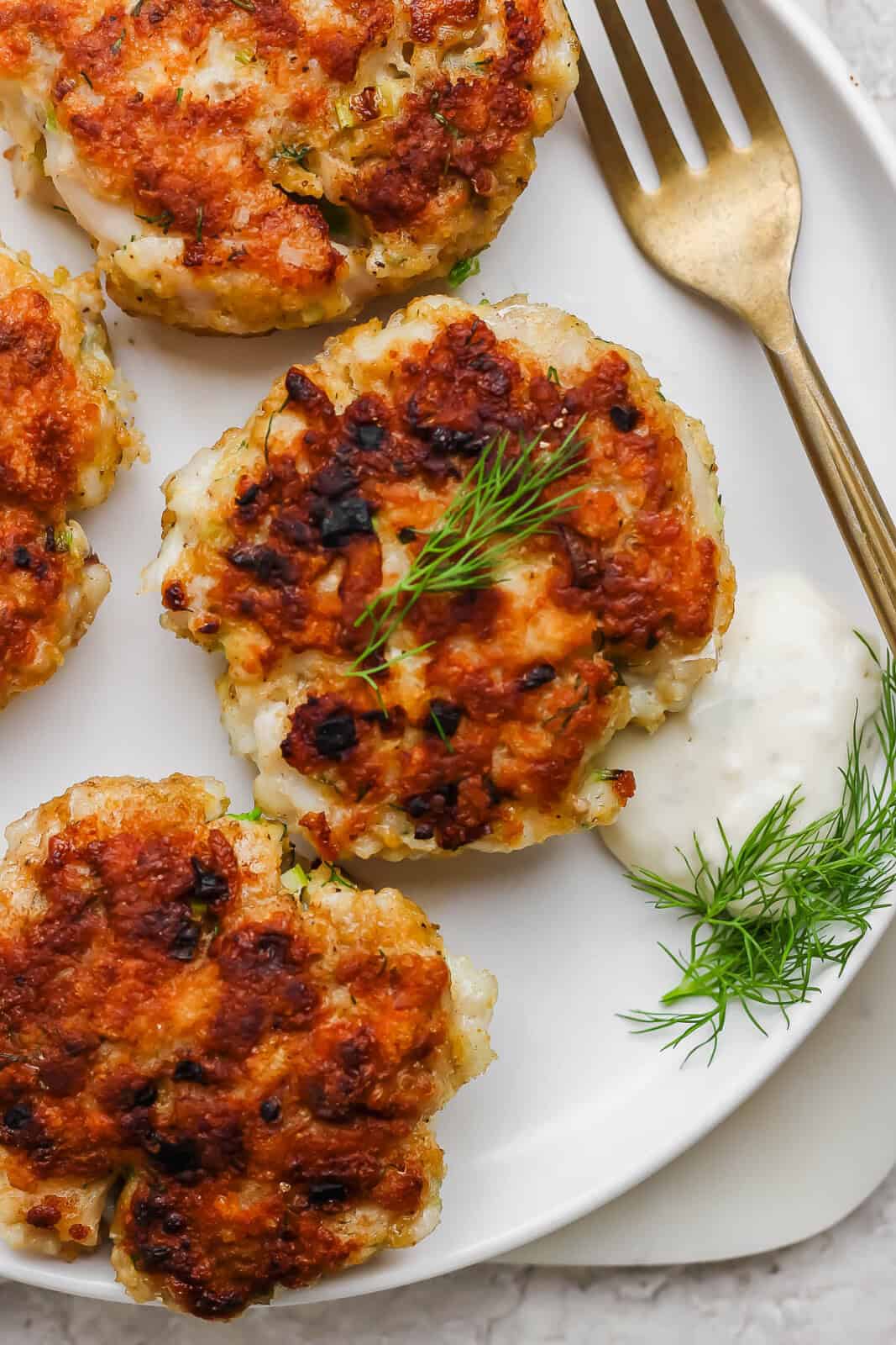 Cooked cod fish cakes on a plate with a fork, fresh dill and tartar sauce.