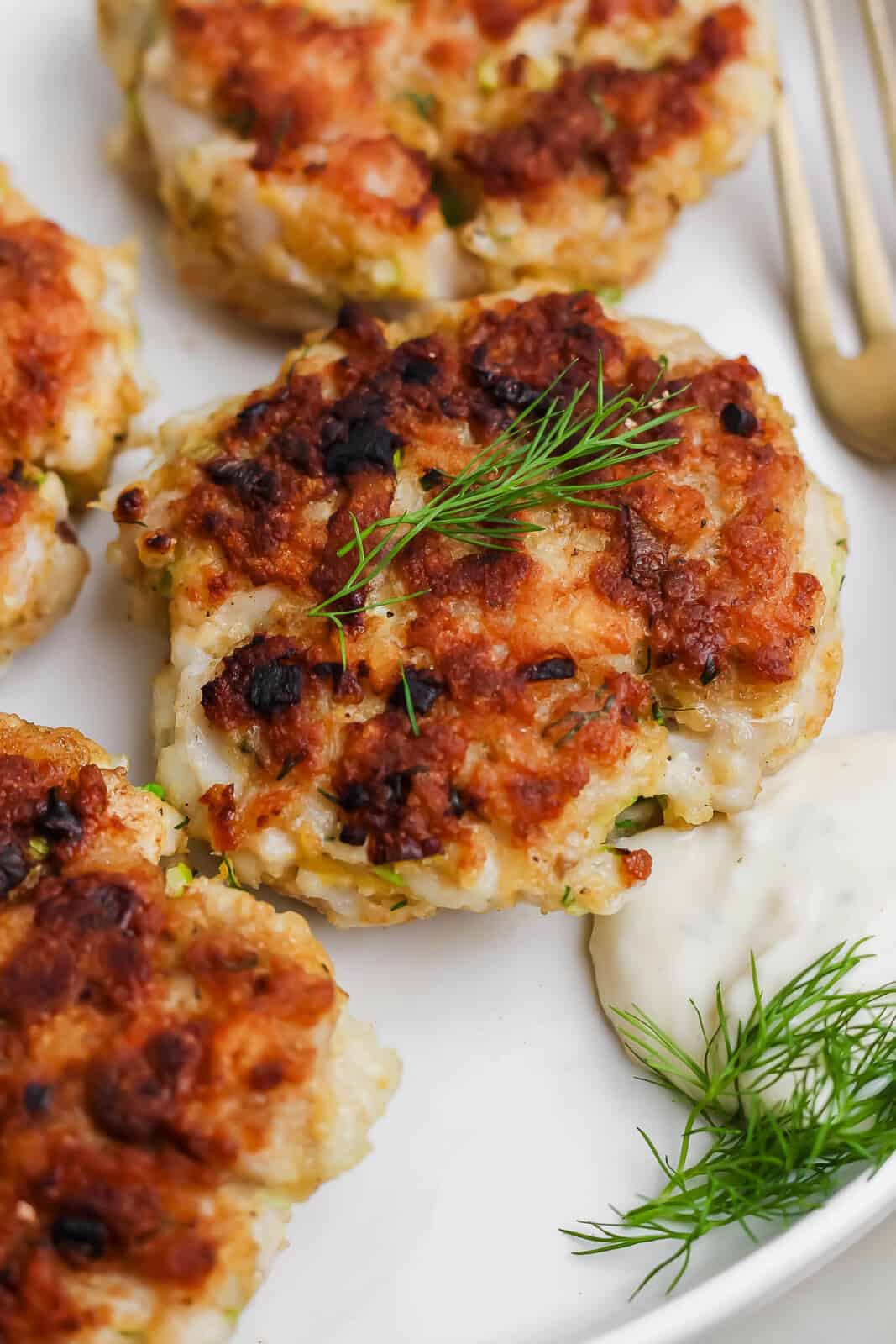 A cooked cod cake a plate with fresh dill on top and tartar sauce next to it.