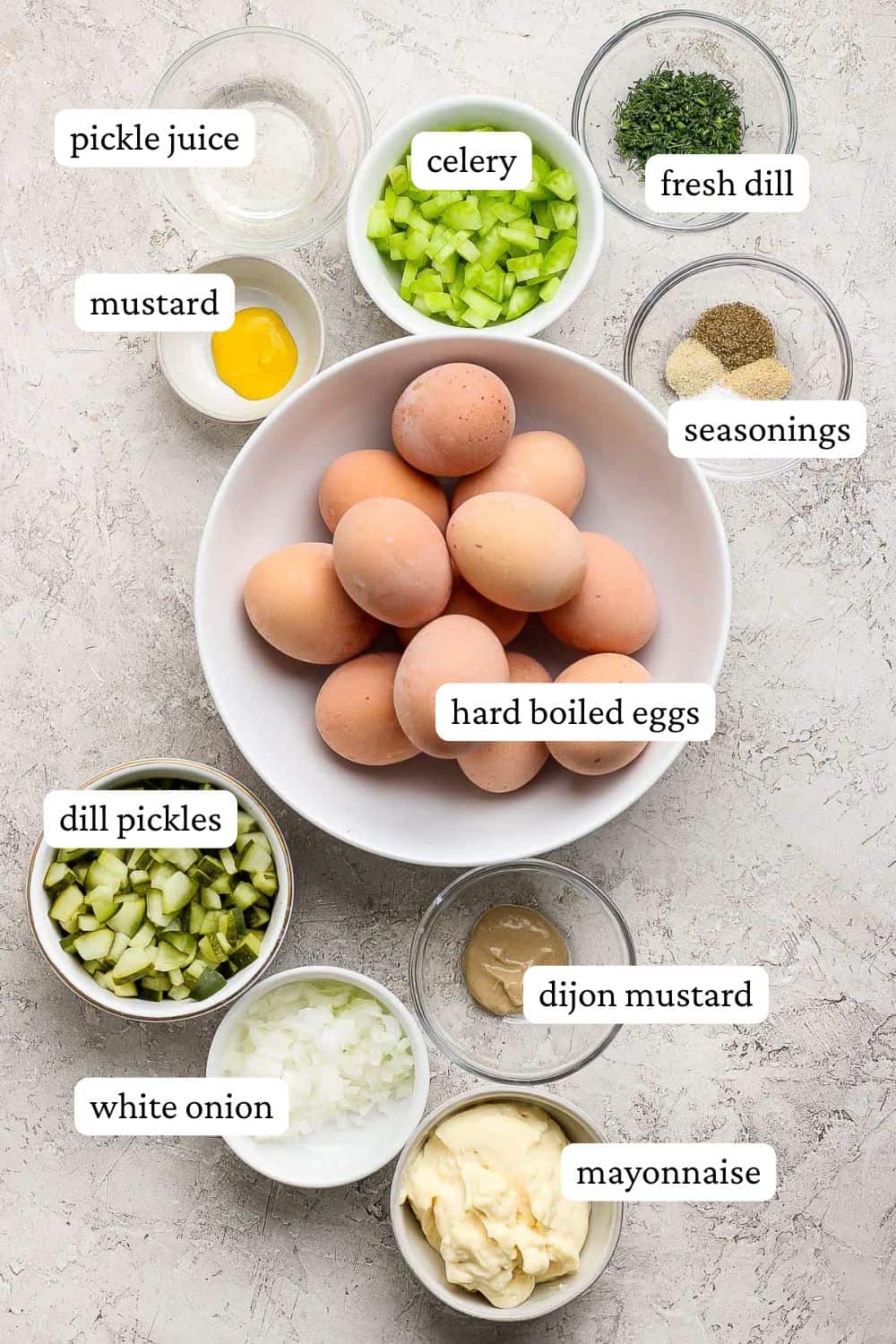 Dill pickle egg salad ingredients in individual bowls; hard boiled eggs, yellow mustard, pickle juice, diced celery, seasonings, dill pickles, dijon mustard, white onion, and mayonnaise.