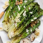 Grilled romaine salad on a platter with Caesar dressing, parmesan cheese and croutons with a wooden knife on the side.