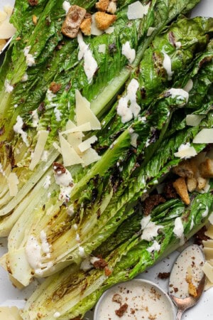 Grilled romaine salad on a platter with Caesar dressing, parmesan cheese and croutons with a wooden knife on the side.