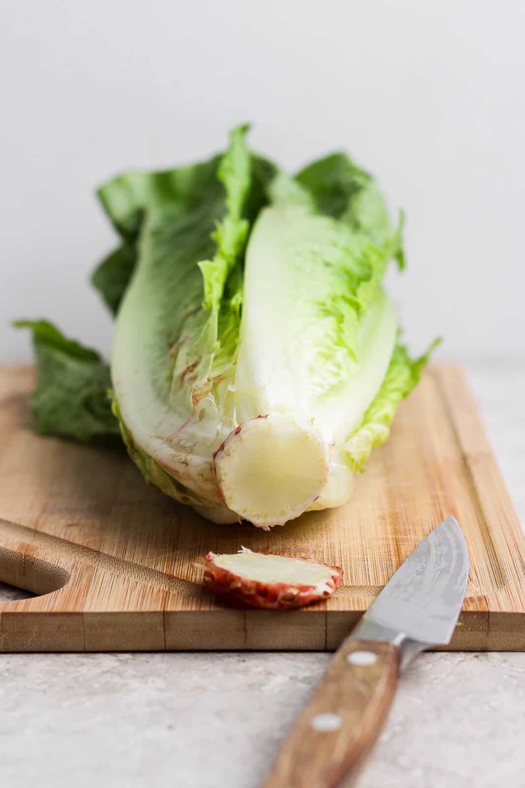 A piece of romaine lettuce on a cutting board with the core facing the camera and a thin slice cut off with a wooden knife laying next to it.