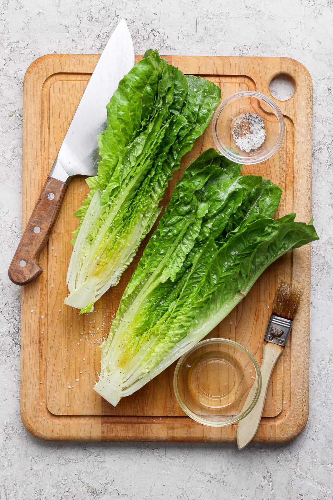 A large wooden cutting board with a large wooden chef's knife, a bowl fo salt and pepper, a bowl of oil and a brush along with two halves of romaine seasoned and brushed with oil.