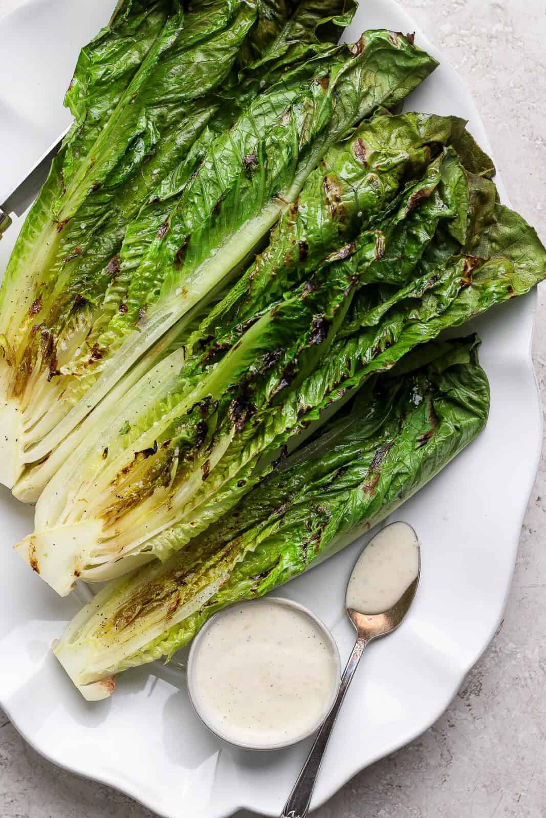 Four pieces of grilled romaine lettuce with char marks on a platter with a bowl of dressing and a spoon.