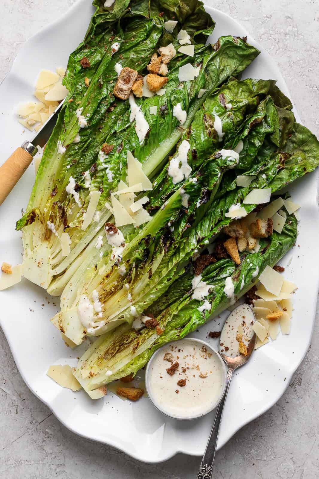 Grilled romaine salad on a platter with Caesar dressing, parmesan cheese and croutons with a wooden knife on the side.
