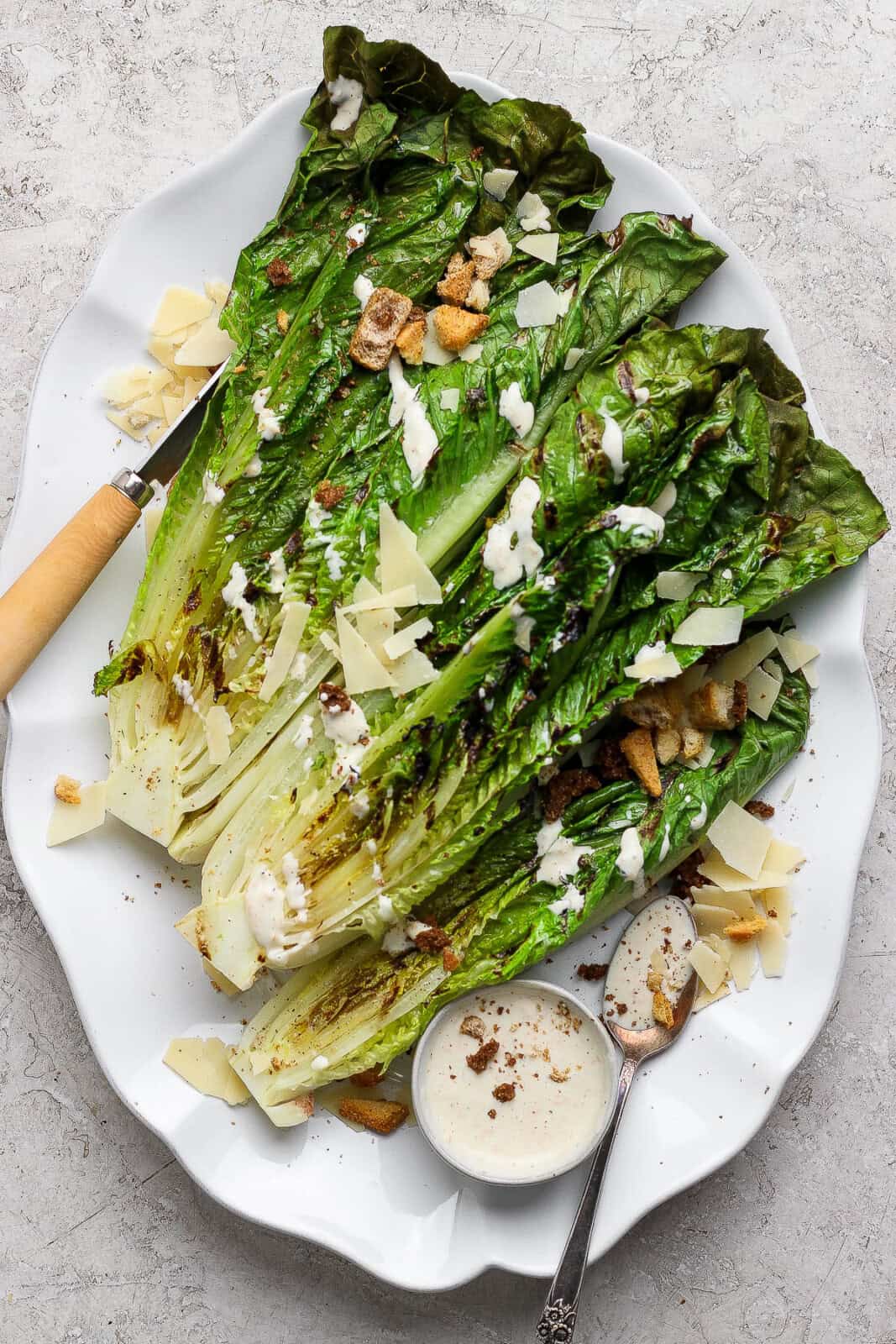 Four pieces of grilled romaine lettuce with char marks on a platter with a bowl of dressing and a spoon. The romaine is dressing with parmesan cheese and croutons.