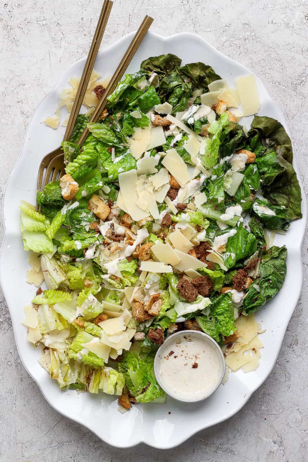 Four pieces of grilled romaine lettuce cut up on a platter with a bowl of dressing and a spoon. Lettuce is dressed with parmesan and croutons.
