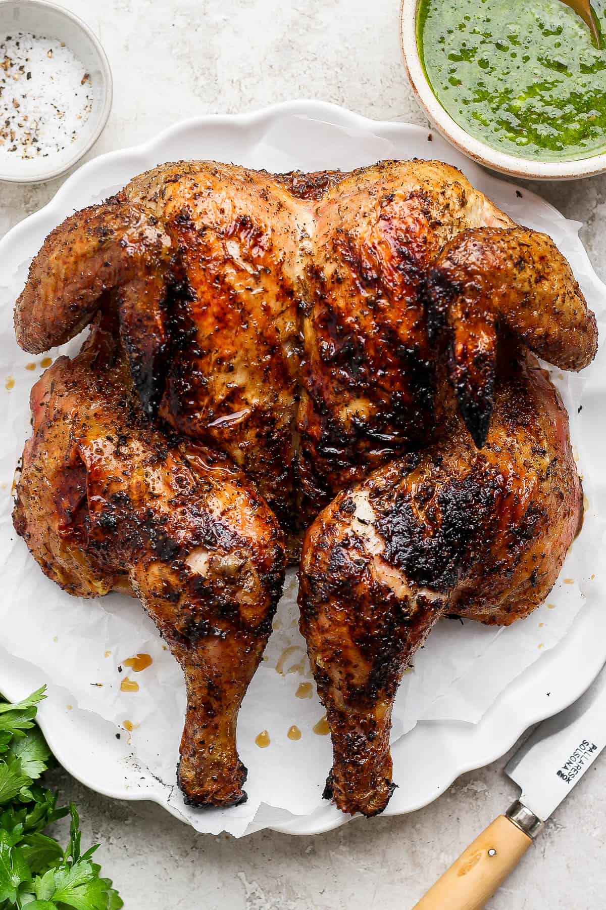 A grilled spatchcock chicken on a platter next to bowls of chimichurri sauce and salt and pepper.