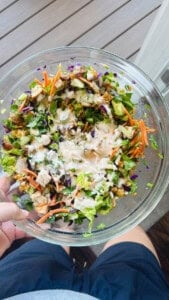 A hand holding a bowl of bang bang chicken salad with dressing on it and she is next to a window.