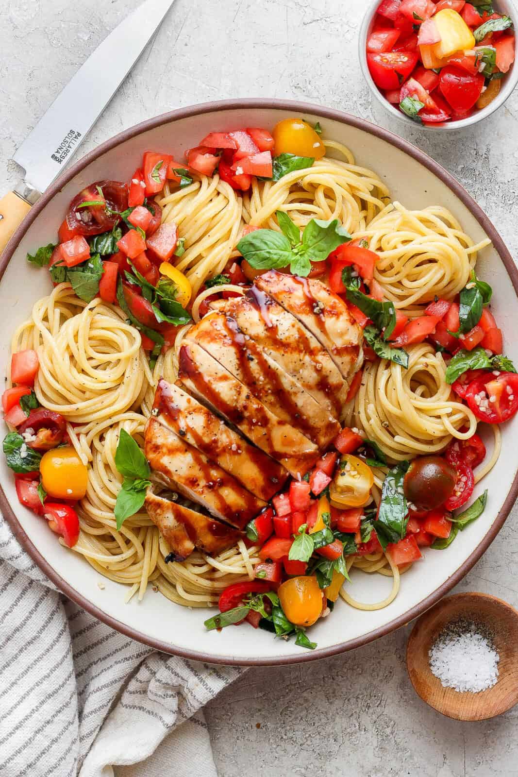 Bruschetta chicken pasta in a large bowl next to a knife and a small bowl of tomatoes.