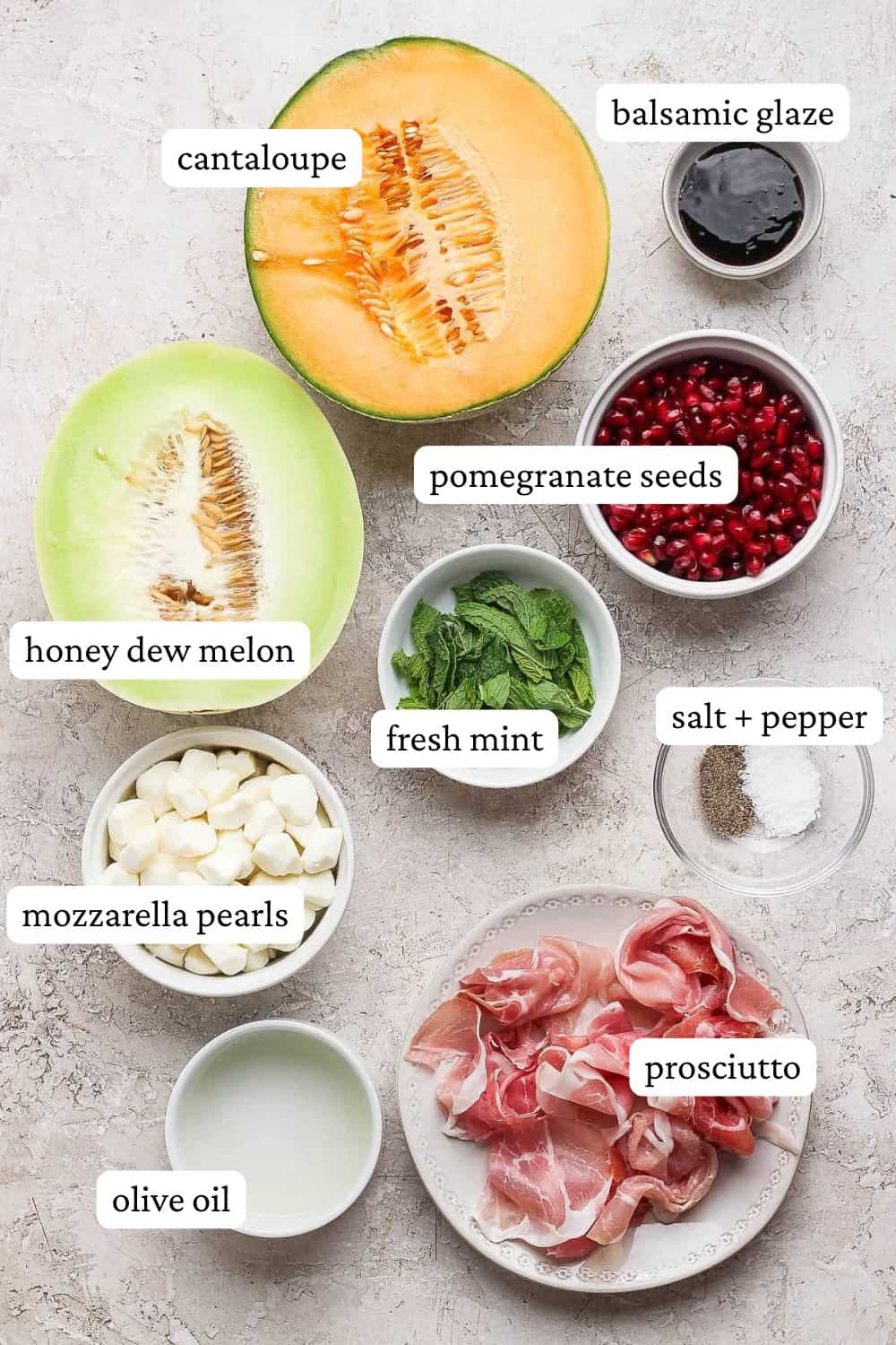 Ingredients for caprese melon salad in small bowls next to melon halves with labels identifying each one.
