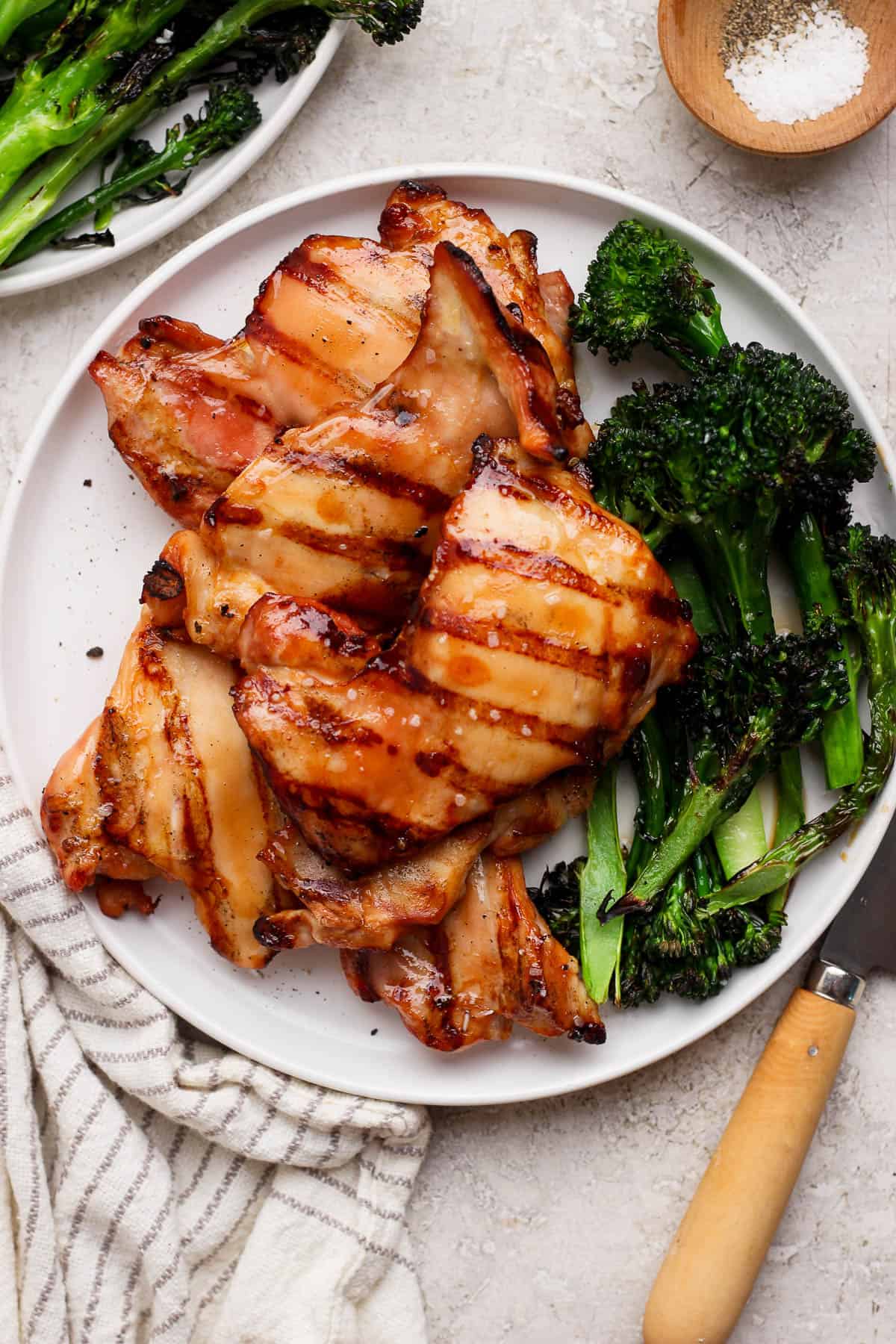 Grilled boneless chicken thighs on a white plate with grilled broccolini next to a knife and small dish of salt and pepper.