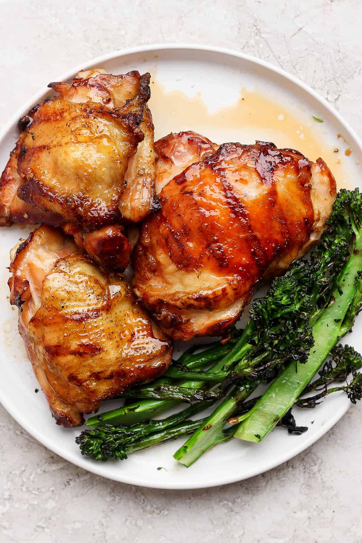 Bone-in grilled chicken thighs on a white plate with grilled broccolini.
