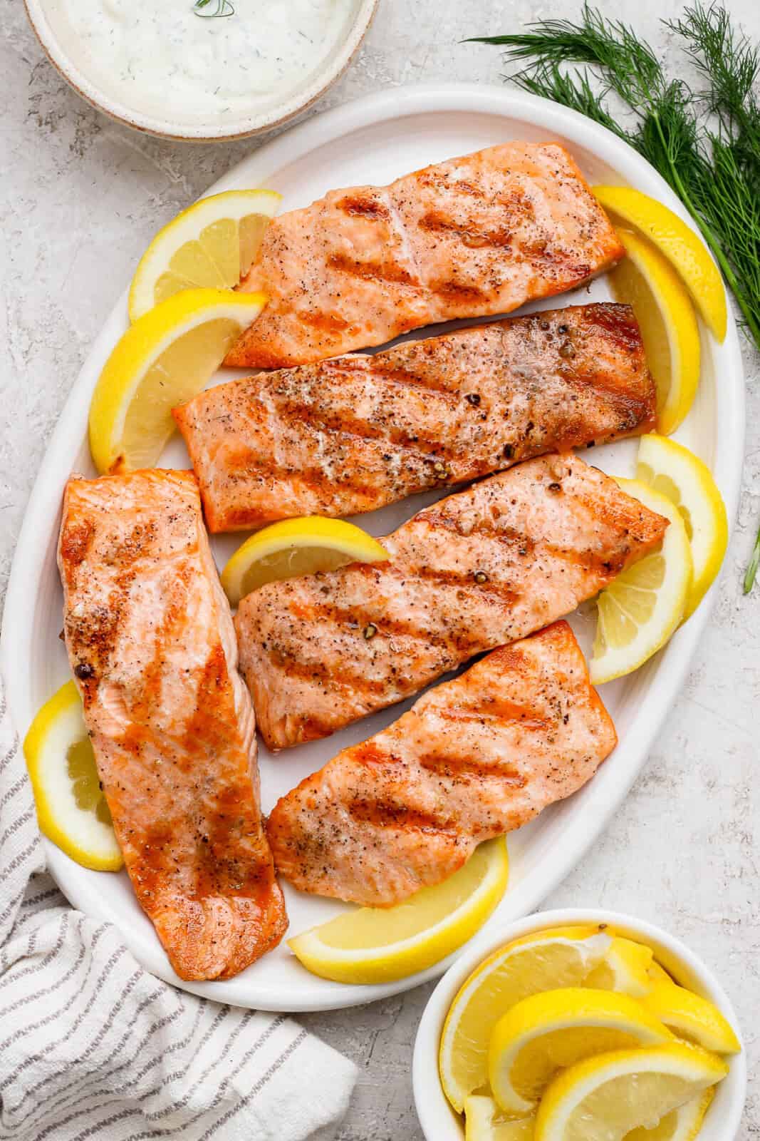 Five grilled salmon fillets on a platter with lemon wedges and fresh dill.