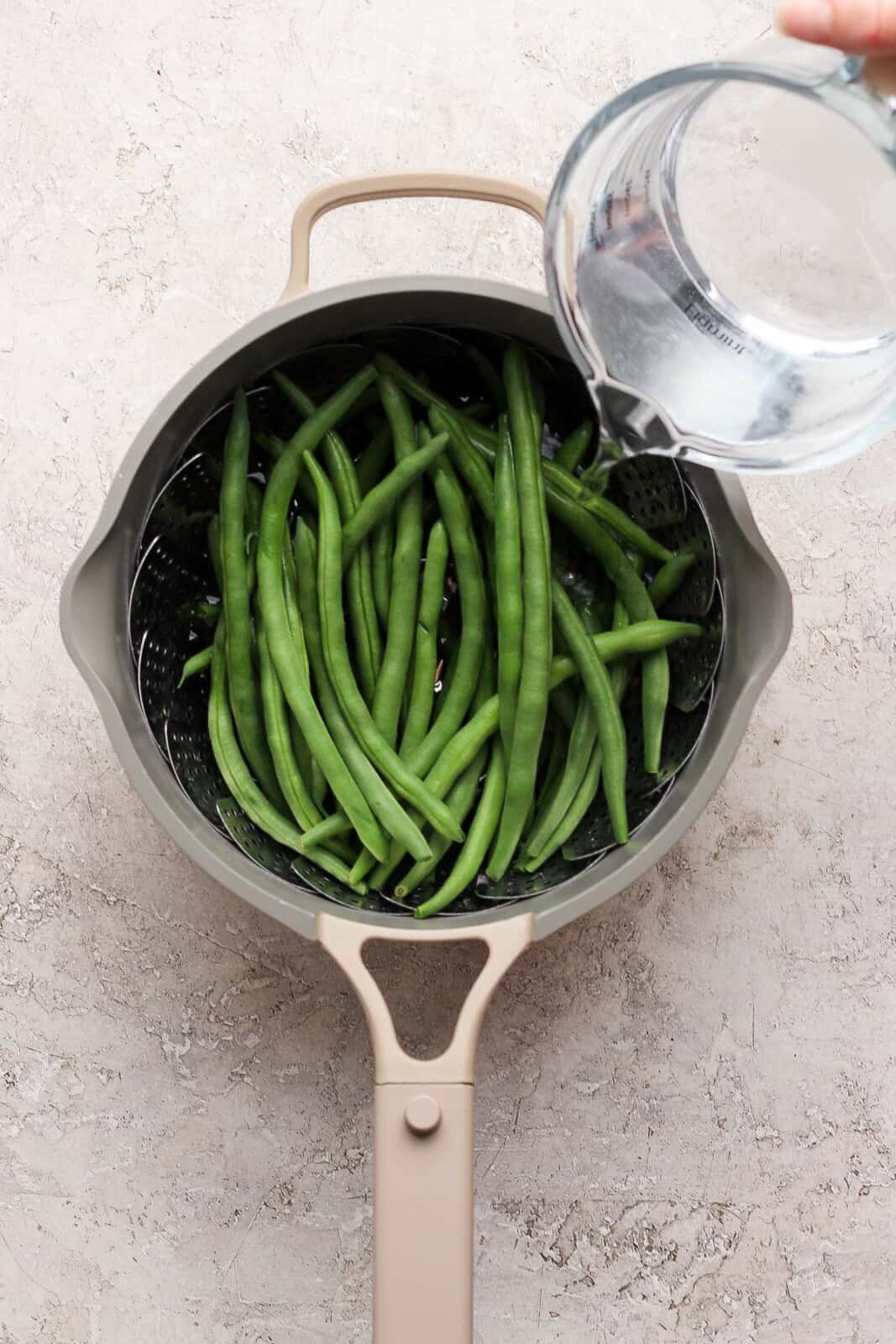 Water being poured into a saucepan with a steamer basket and fresh green beans to make steamed green beans.