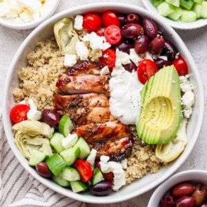 A fully assembled mediterranean chicken quinoa bowl next to small bowls of cucumber, feta, and olives.