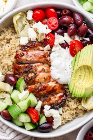 A fully assembled mediterranean chicken quinoa bowl next to small bowls of cucumber, feta, and olives.