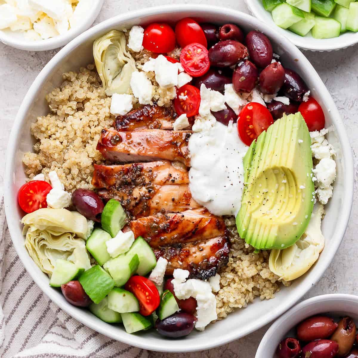 A fully assembled mediterranean chicken quinoa bowl next to small bowls of cucumber, feta, and olives.
