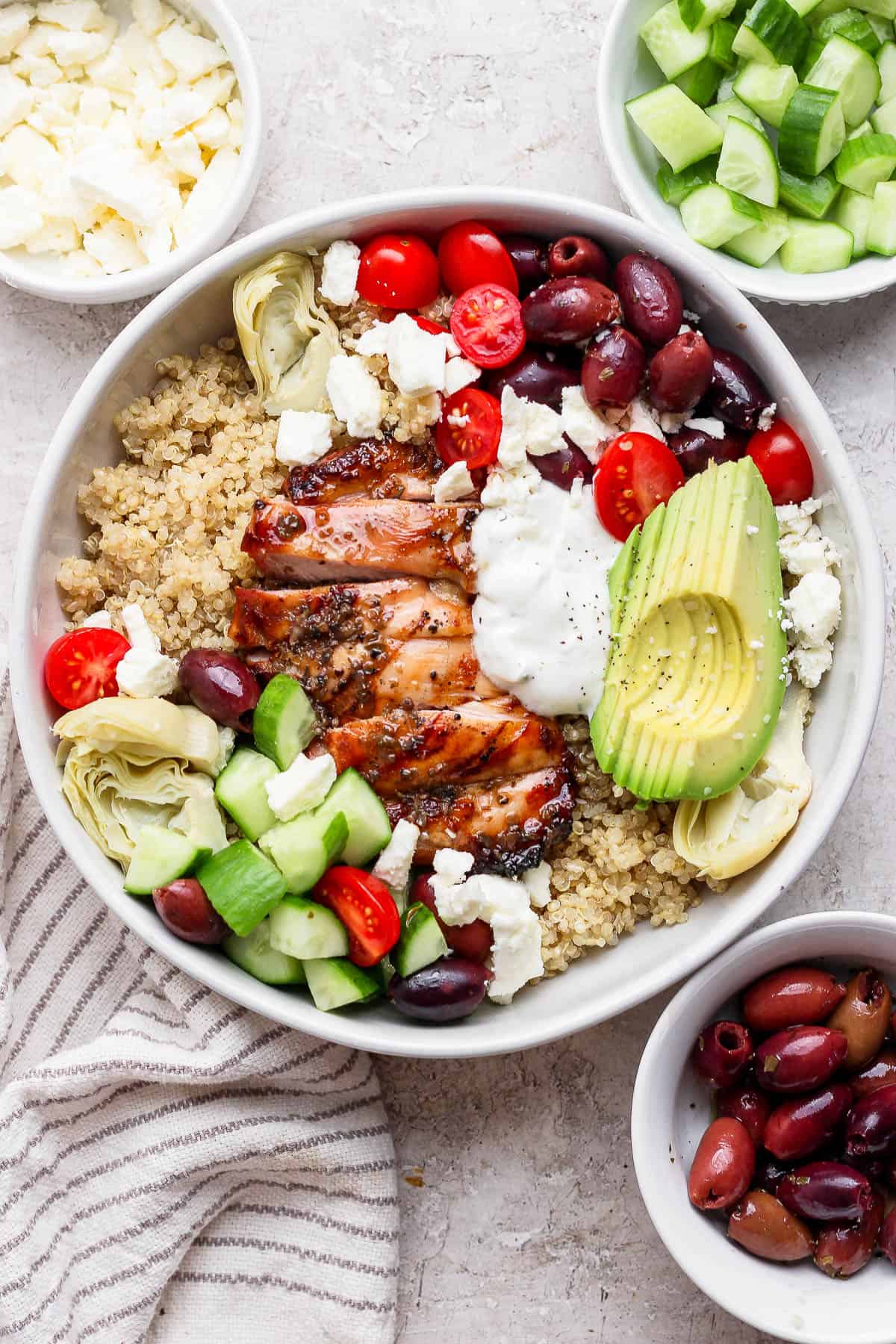A Mediterranean chicken quinoa bowl next to small bowls of cucumbers, feta, and olives.