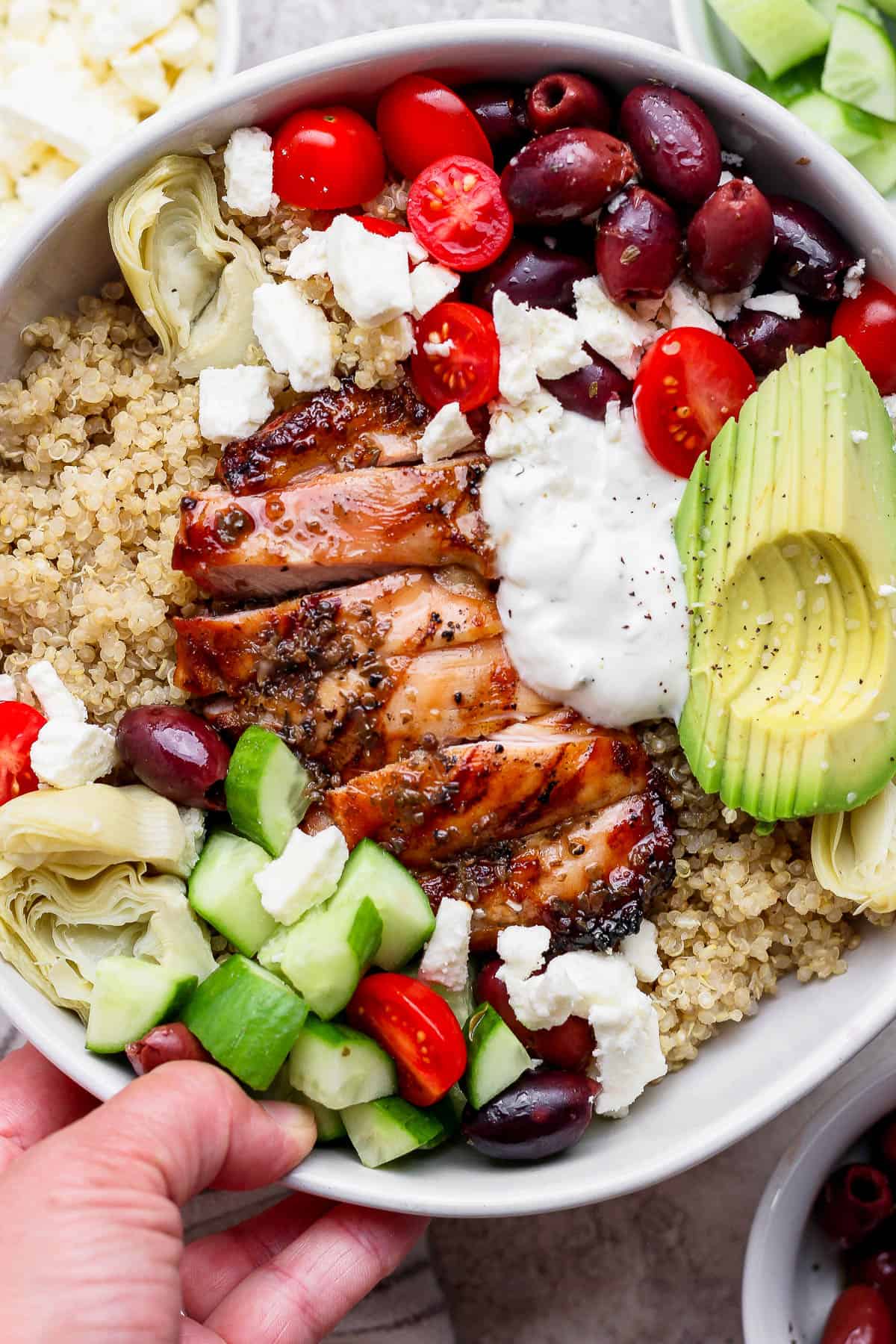 A hand holding a fully assembled Mediterranean chicken quinoa bowl.
