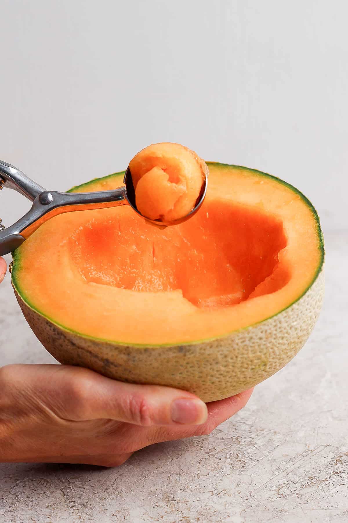 Someone scooping a ball of cantaloupe out of one half of the melon for a caprese melon salad.