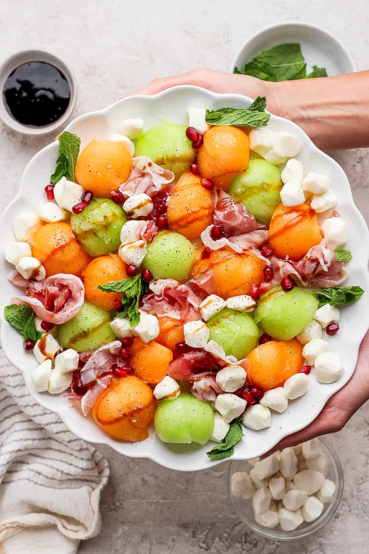 Two hands holding a caprese melon salad next to small bowls of mozzarella and balsamic glaze.