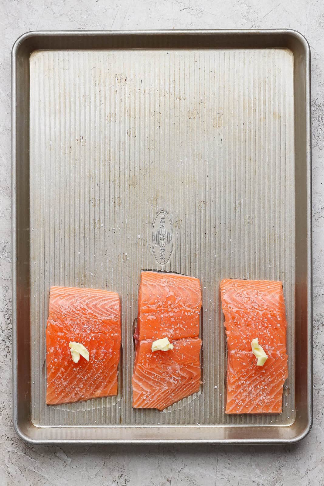 Three raw salmon fillets with dabs of butter on top on one side of a sheet pan to make salmon power bowls.