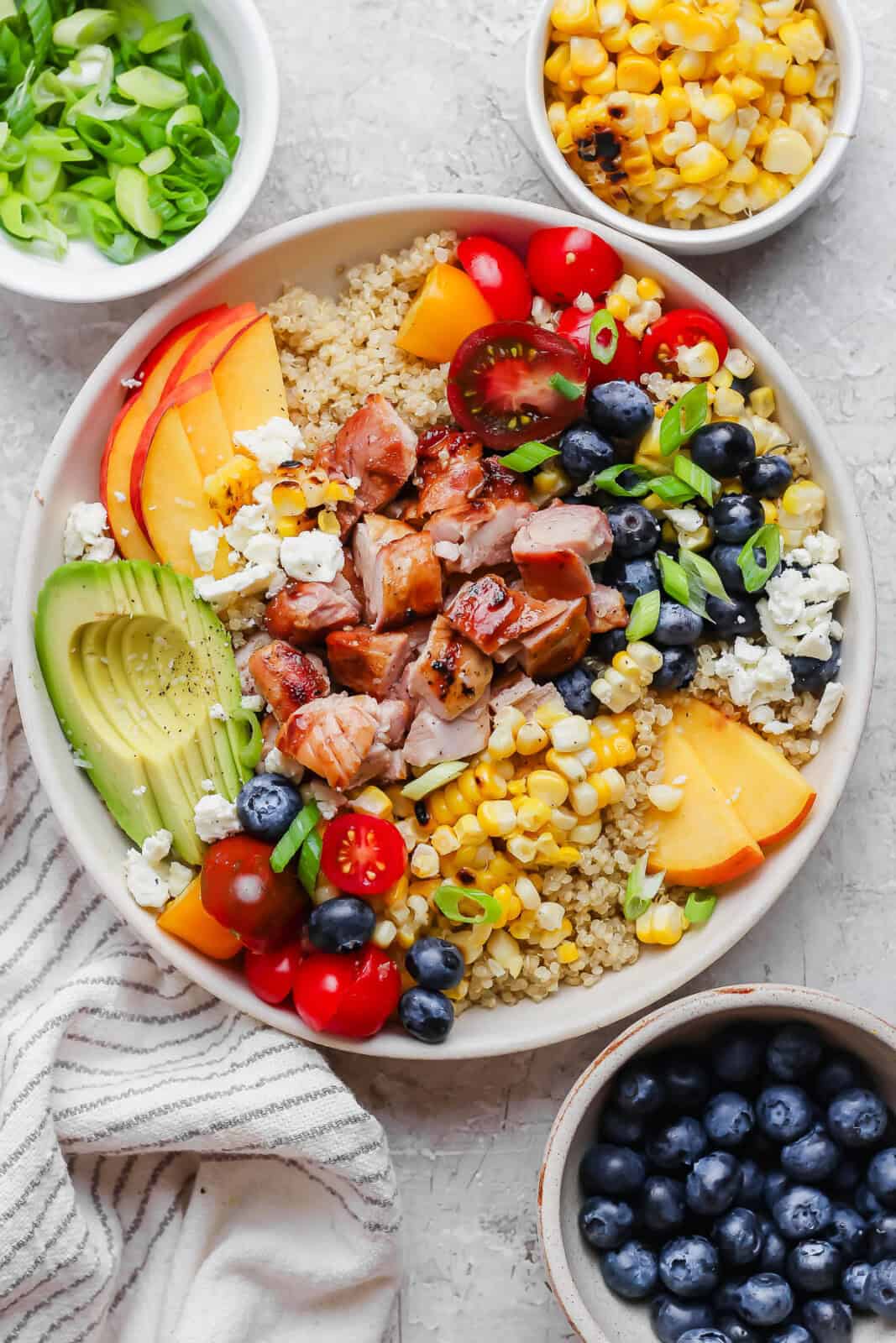 A fully assembled summer quinoa salad bowl next to small bowls of green onions, corn kernels, and blueberries.