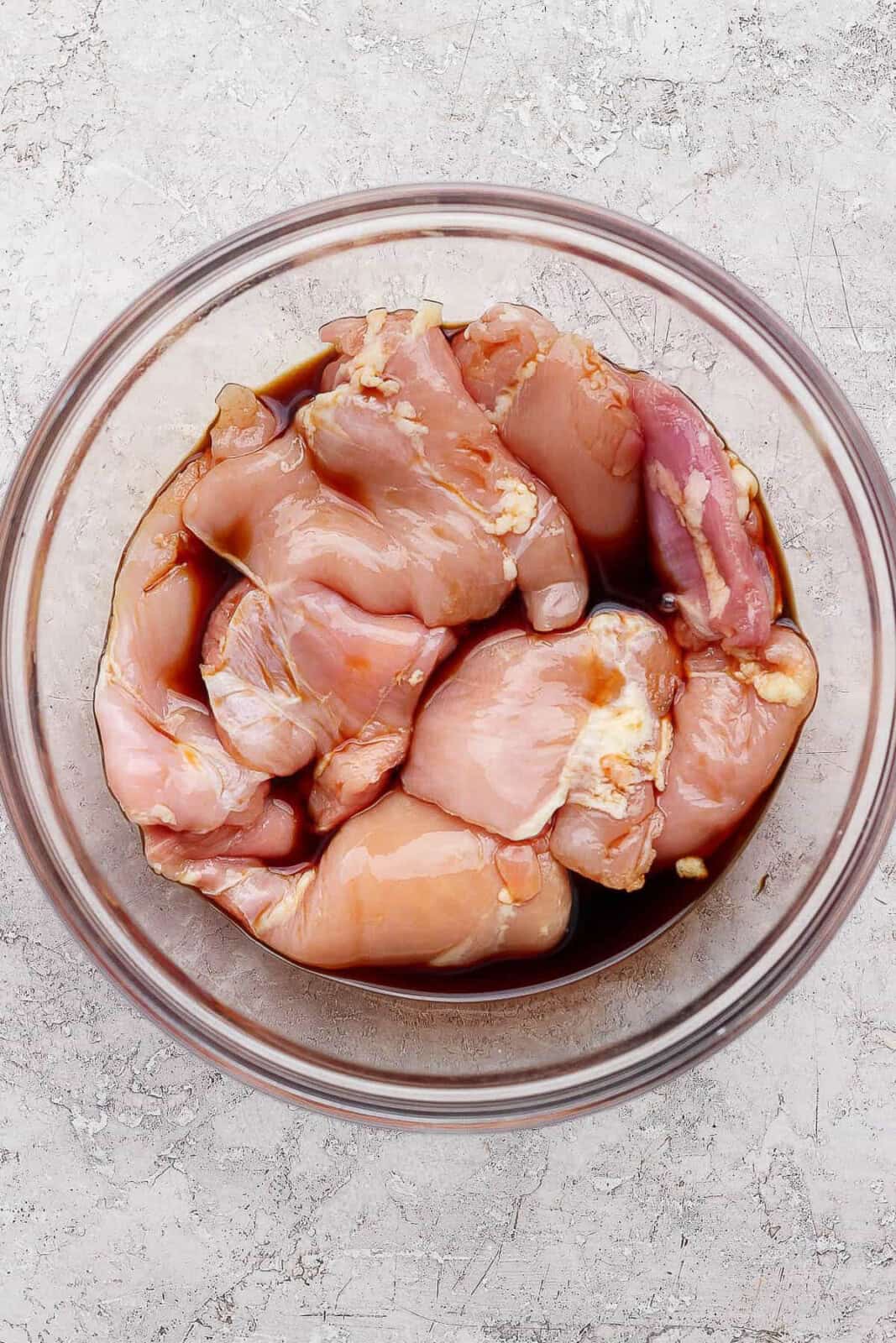 Raw chicken thighs marinating in soy sauce in a large glass bowl.