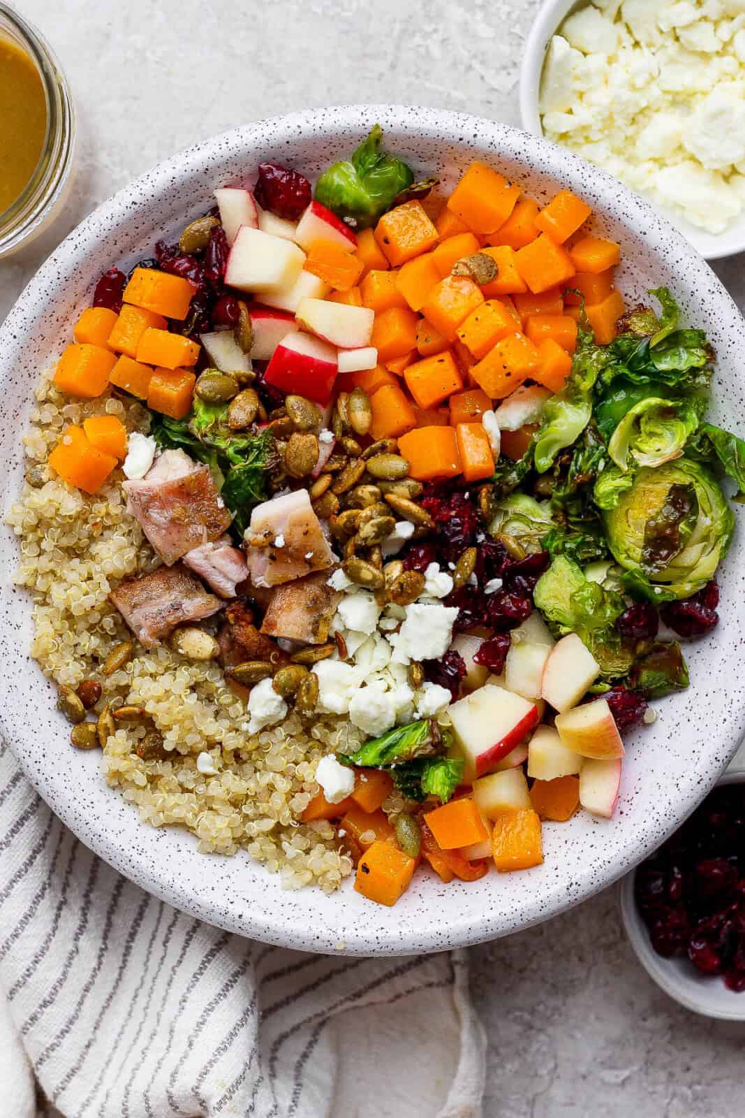 A fully assembled Autumn harvest bowl next to small bowls of dressing, feta cheese, and dried cranberries.