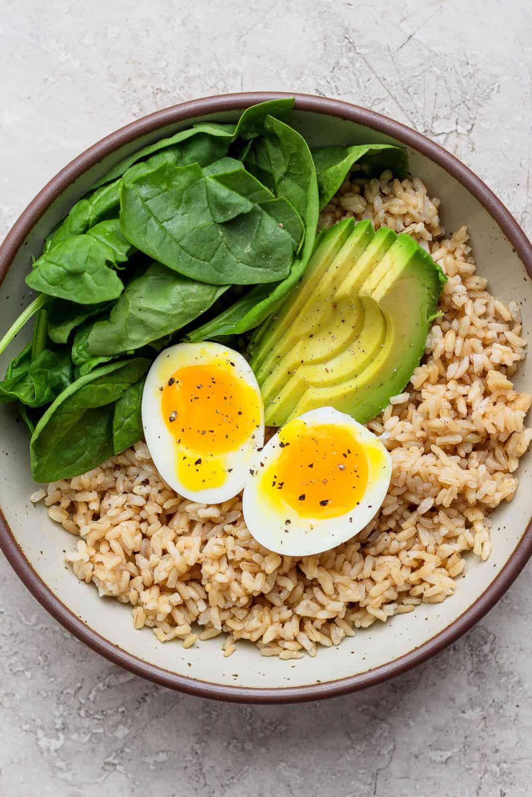 An avocado egg bowl with brown rice, spinach, sliced avocado, and a soft boiled egg cut in half.