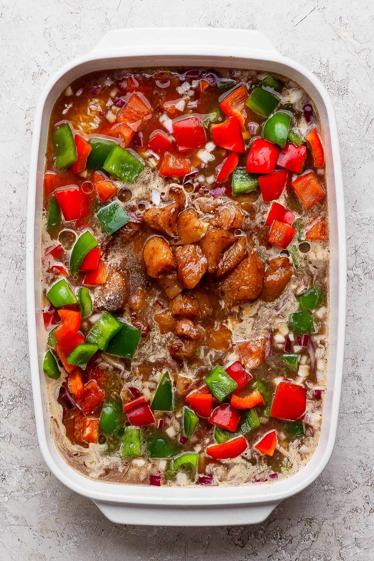 Chopped bell pepper, diced red onion, white rice, chicken broth, olive oil, lime zest, and fajita seasoning added to the casserole dish to make chicken fajita casserole with rice.
