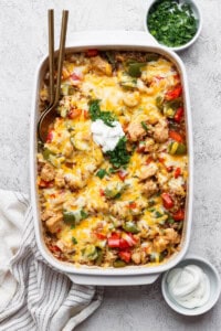 A fully baked chicken fajita casserole with rice in a white baking dish next to small bowls of cilantro and sour cream.