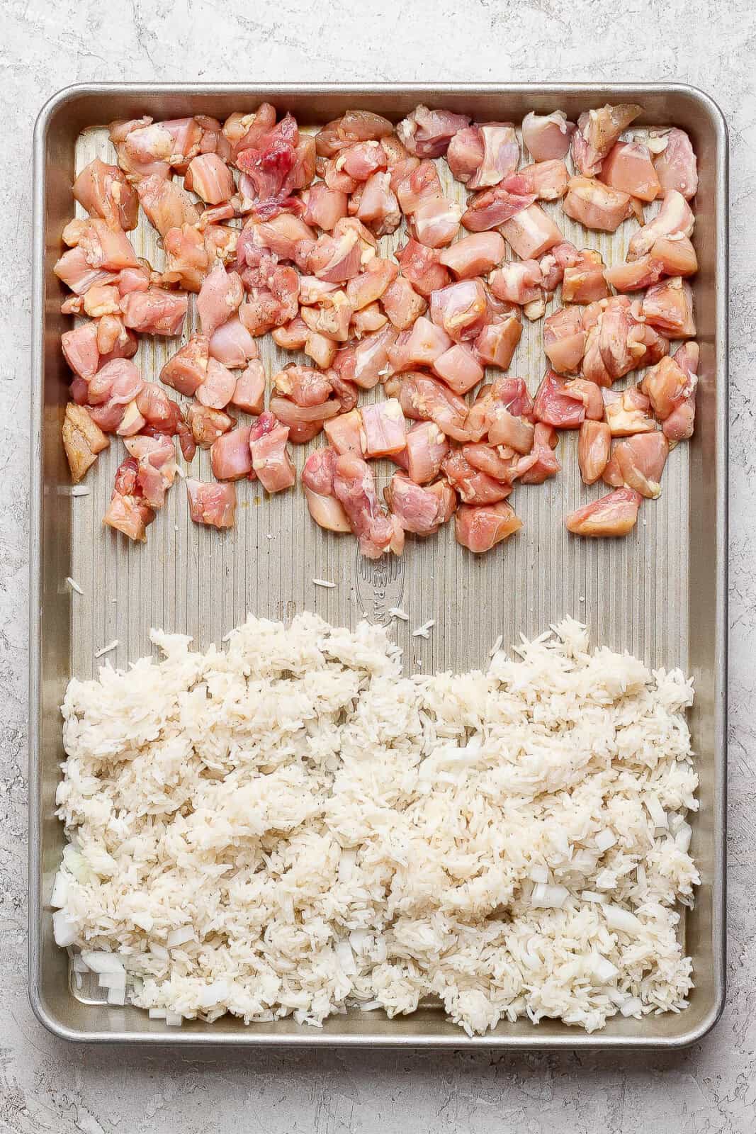 Raw cut-up chicken thighs on one side of a sheet pan and rice with onions on the other side before cooking into sheet pan fried rice.