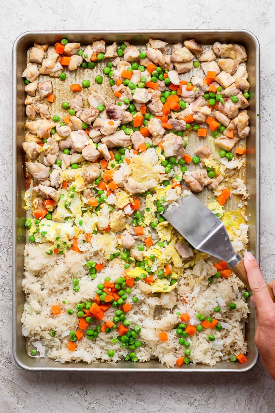 A spatula spraping the cooked eggs from the bottom of the pan to toss with the rest of the sheet pan fried rice ingredients.