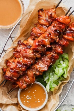 Thai chicken skewers in a basket with a peanut dip and marinated cucumbers.