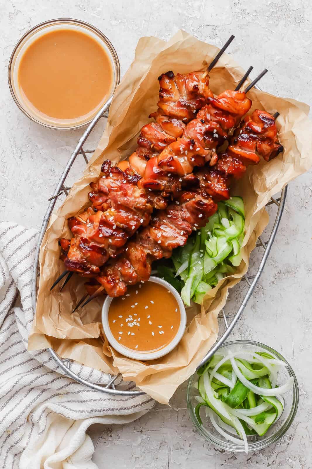 Thai chicken skewers in a basket lined with parchment paper and a dish of peanut sauce next to some marinated cucumbers.