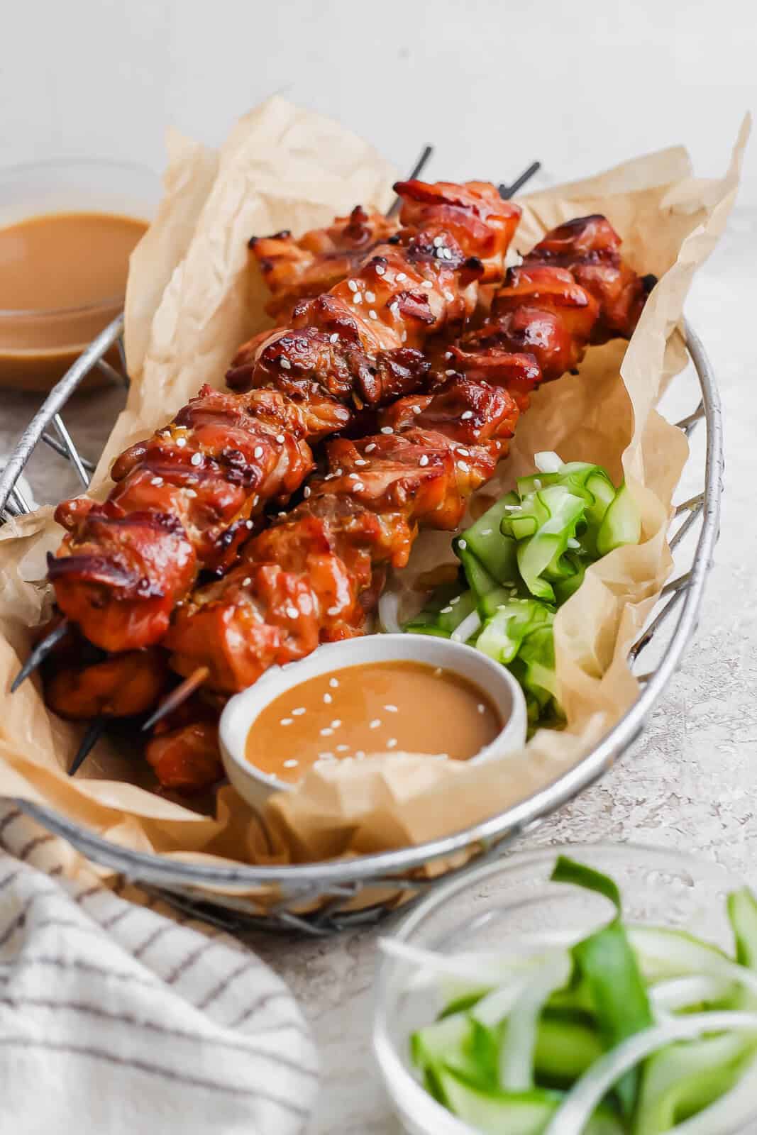 Thai chicken skewers in a basket with marinated cucumbers and a dish of peanut sauce dip.