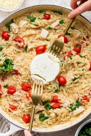 Boursin orzo with chicken in a large braiser with the Boursin cheese in the middle being pulled apart with two forks.