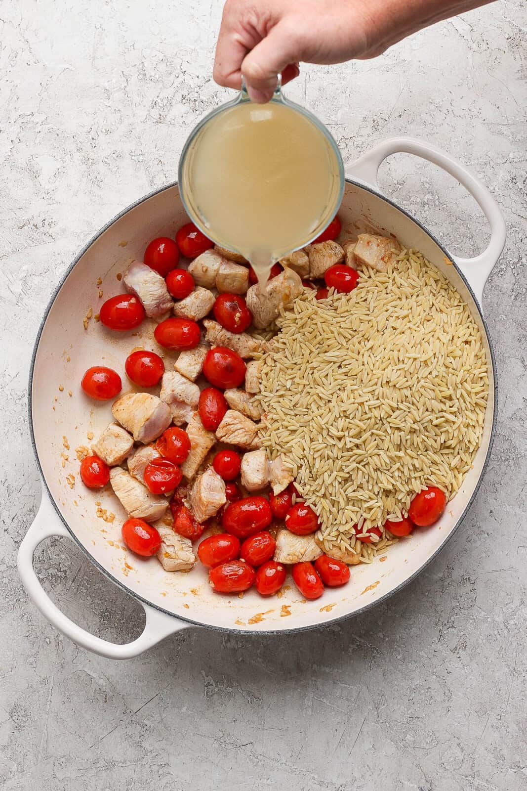 Chicken broth being poured into the pan of seared chicken, tomatoes, and orzo for making boursin orzo with chicken.