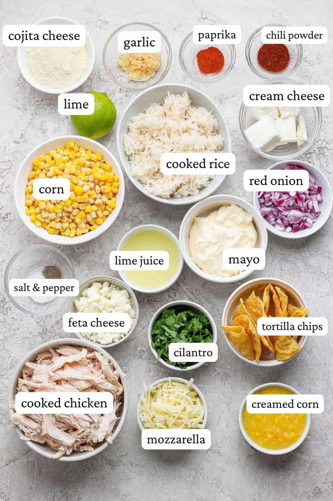 Street corn chicken casserole ingredients in individual bowls - cojita cheese, garlic, paprika, chili powder, lime, cooked rice, cream cheese, corn, salt & pepper, lime juice, mayo, red onion, feta cheese, cilantro, tortilla chips, cooked chicken, mozzarella cheese, and creamed corn.